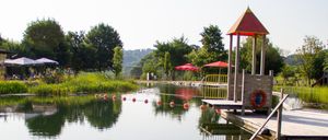 Ein ruhiger Wasserpark mit einem kleinen Spielplatz, umgeben von üppiger Grünfläche und Bäumen. Das Wasser ist ruhig, mit roten und weißen Bojen, die einen sicheren Schwimmbereich markieren.