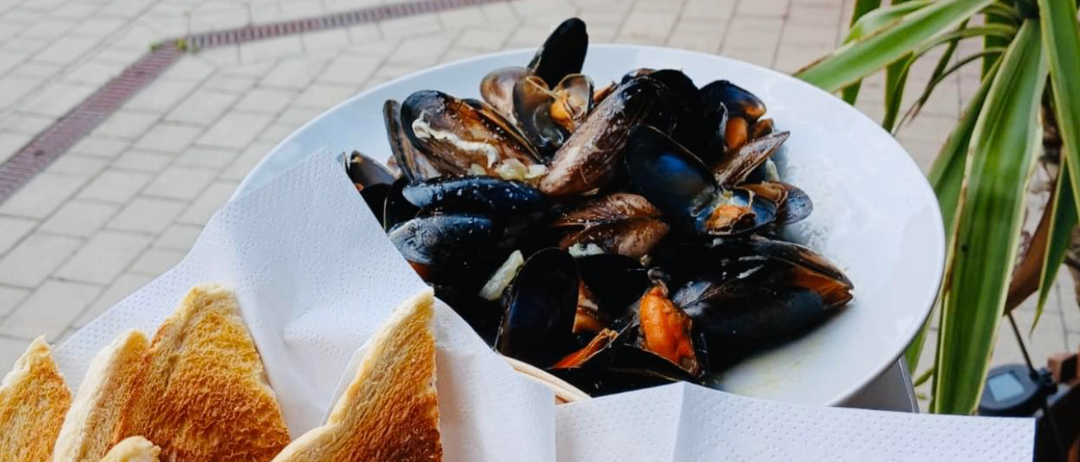Ein weißer Teller mit Muscheln in einer Brühe, serviert mit geröstetes Brot auf der Seite, auf einem Ziegelboden platziert.