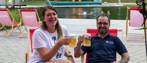 Ein Mann und eine Frau sitzen auf Stühlen in der Nähe eines Pools, lächeln und halten Gläser mit Bier in die Höhe. Dahinter schwimmen aufblasbare Spielzeuge im Wasser, und ein Rasenbereich mit Bäumen ist sichtbar.