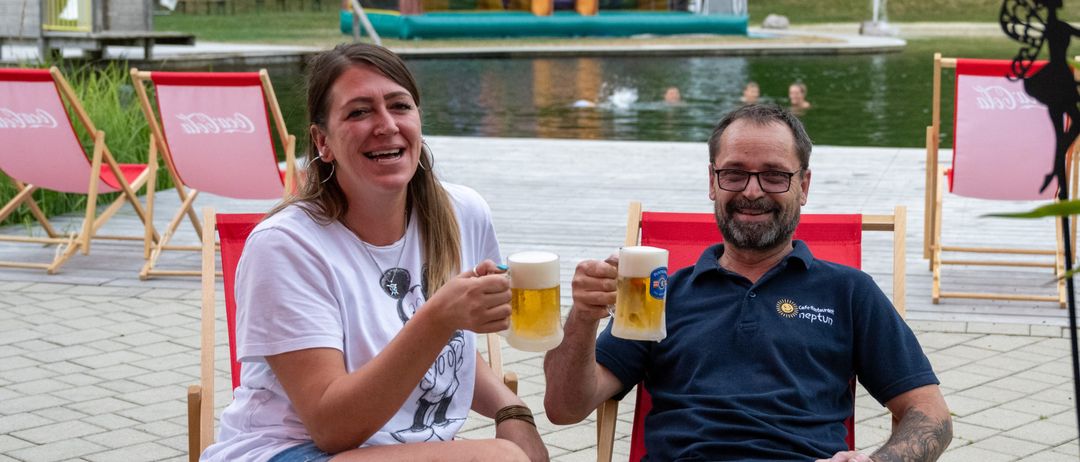 Ein Mann und eine Frau sitzen auf Stühlen in der Nähe eines Pools, lächeln und halten Gläser mit Bier in die Höhe. Dahinter schwimmen aufblasbare Spielzeuge im Wasser, und ein Rasenbereich mit Bäumen ist sichtbar.