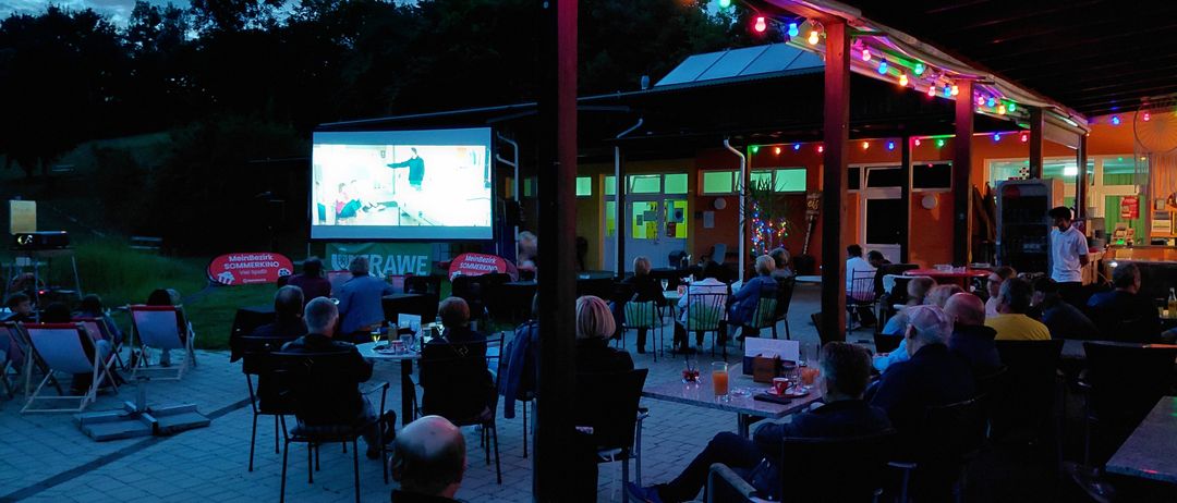 Menschen versammeln sich bei einem Open-Air-Kino und schauen einen Film. Der Bereich ist mit farbigen Lichtern beleuchtet.