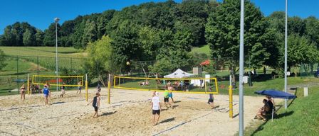 Mehrere Teams spielen Volleyball auf einem sandigen Platz umgeben von grünem Gras. Bäume und ein weißes Zelt im Hintergrund.