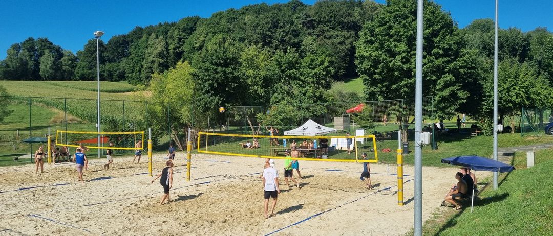 Mehrere Teams spielen Volleyball auf einem sandigen Platz umgeben von grünem Gras. Bäume und ein weißes Zelt im Hintergrund.
