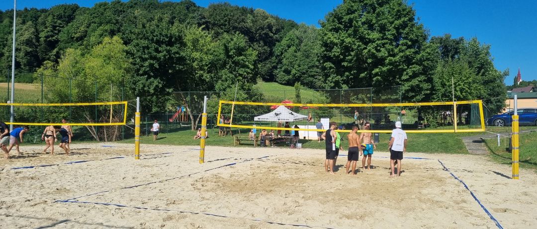 Mehrere Personen spielen Beachvolleyball auf einem Sandplatz. Der Platz hat gelbe Netze. Menschen stehen herum und sitzen auf Bänken. Bäume und ein Spielplatz sind im Hintergrund.