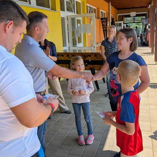 Mehrere Erwachsene und Kinder stehen in einem Flur und schütteln sich die Hände. Ein junges Mädchen hält einen Becher und einen Jungen eine Karte. Ein anderes Mädchen trägt eine Brille und ein blaues Shirt.