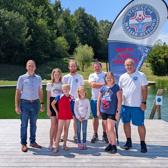 Sechs Personen posieren für ein Foto auf einem Steg am Wasser, mit einem Banner hinter ihnen. Das Banner steht 'Notruf über LWW00'.