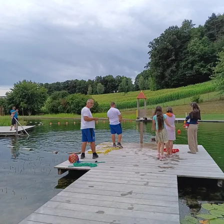 Mehrere Menschen versammeln sich auf einem Holzsteg über einem See, einige halten Angelruten. Zwei Männer stehen auf dem Steg, während andere am Ufer des Wassers stehen. Bäume und ein Feld sind im Hintergrund zu sehen.