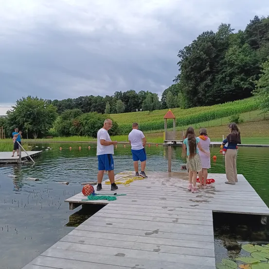 Mehrere Menschen versammeln sich auf einem Holzsteg über einem See, einige halten Angelruten. Zwei Männer stehen auf dem Steg, während andere am Ufer des Wassers stehen. Bäume und ein Feld sind im Hintergrund zu sehen.