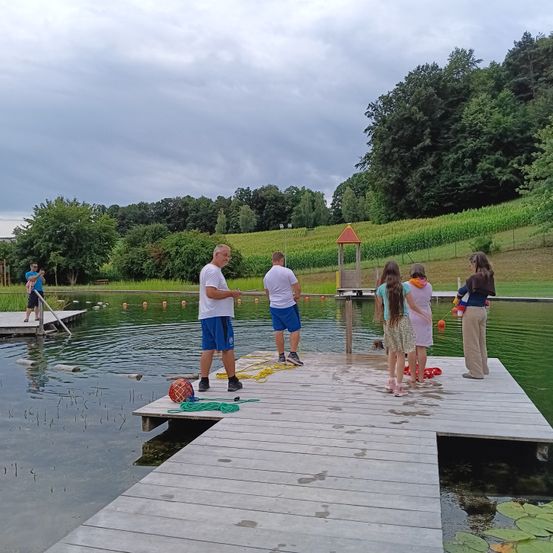 Mehrere Menschen versammeln sich auf einem Holzsteg über einem See, einige halten Angelruten. Zwei Männer stehen auf dem Steg, während andere am Ufer des Wassers stehen. Bäume und ein Feld sind im Hintergrund zu sehen.