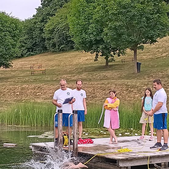 Fünf Personen stehen auf einem Steg in der Nähe des Wassers. Eine Person schwimmt. Zwei Männer halten Klemmbretter. Zwei Frauen und ein Mädchen stehen auf dem Steg. Ein Baum und Bänke stehen hinter ihnen.
