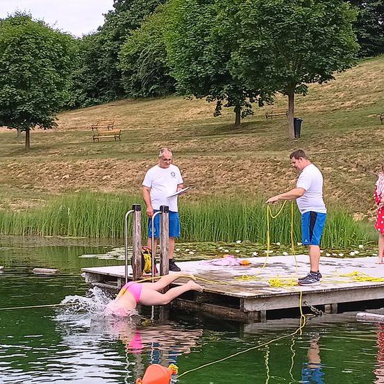 Zwei Männer stehen auf einem Steg, einer hält ein Seil. Eine Frau in einem pinken Badeanzug taucht ins Wasser. Dahinter befindet sich ein Grasbereich mit Bäumen und Bänken.