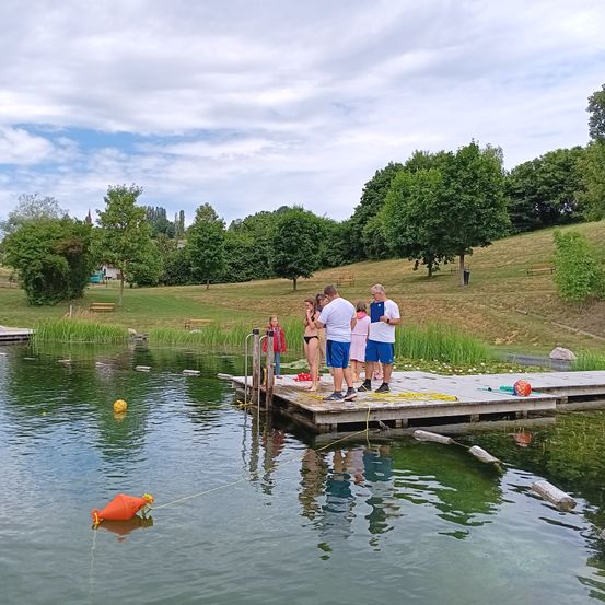 Eine Gruppe von Menschen in Badekleidung steht auf einem Holzsteg und bereitet sich darauf vor, ins Wasser zu springen. Bäume und ein grasbewachsenes Feld umgeben den See. Ein strahlend blauer Himmel mit Wolken ist über ihnen.