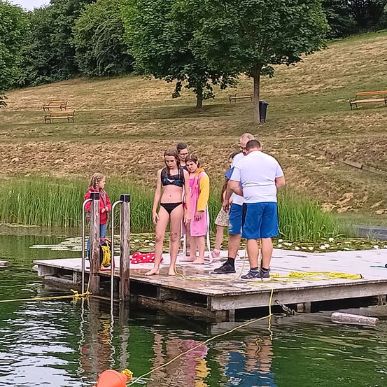 Eine Gruppe von Menschen steht auf einem Steg am See, darunter eine Frau in einem Bikini. Sie sind von Wasser und einer grasbewachsenen Anhöhe umgeben.
