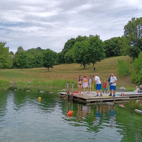 Eine Gruppe von Menschen steht auf einem Steg in der Nähe eines Sees, zwei Männer halten Seile und eine Frau steht in der Nähe eines roten Bojen.