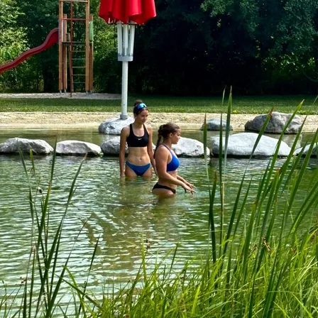 Zwei Frauen in Badekleidung stehen in einem See, umgeben von Felsen und Gras. Hinter ihnen sind ein roter Rutschen und ein roter Schirm zu sehen.