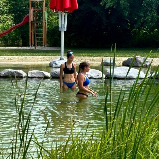 Zwei Frauen in Badekleidung stehen in einem See, umgeben von Felsen und Gras. Hinter ihnen sind ein roter Rutschen und ein roter Schirm zu sehen.