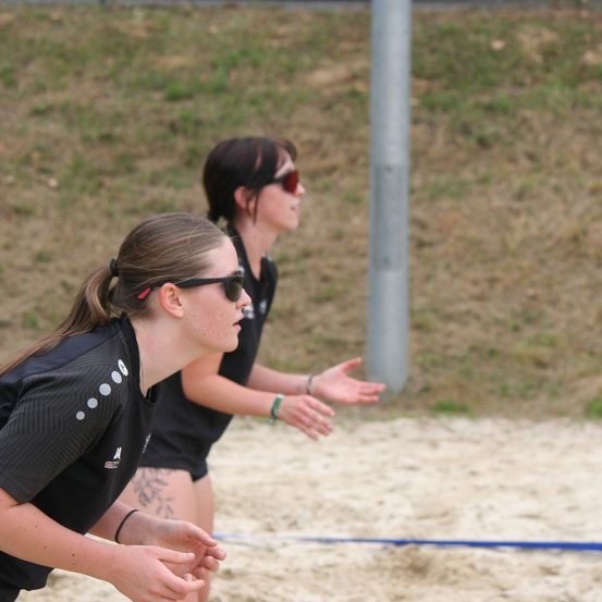 Zwei Frauen in schwarzen Trikots und Sonnenbrillen spielen Volleyball auf einem sandigen Feld. Eine Frau schaut nach links, die andere nach rechts, mit einem Pfahl hinter ihnen.