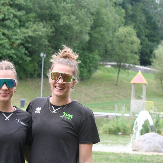 Zwei Frauen in schwarzen Shirts mit weißen Logos stehen eng beieinander, lächeln und posieren für ein Foto. Sie tragen Sonnenbrillen und stehen vor einem Park mit einem Teich.