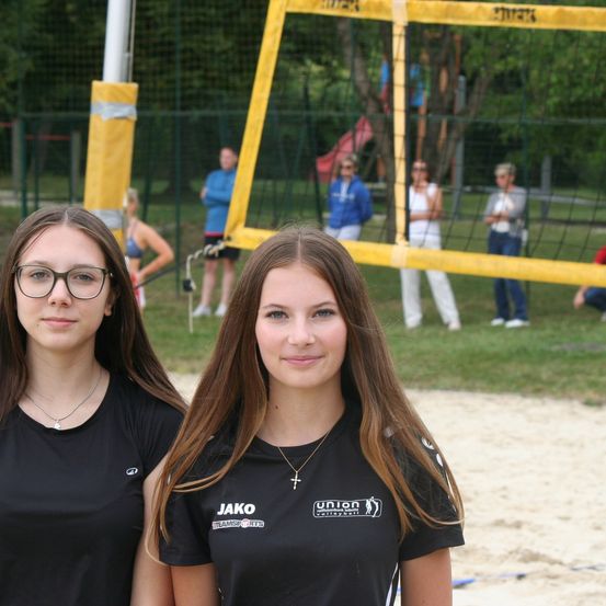 Zwei junge Frauen stehen lächelnd vor einem Beachvolleyballnetz. Andere Personen sind im Hintergrund zu sehen, in der Nähe des Netzes und auf der Wiese.