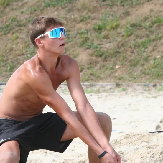 Ein junger Mann ist am Strand, trägt eine Sonnenbrille und schwarze Shorts, mit ausgestreckten Armen und kniet auf dem Sand.