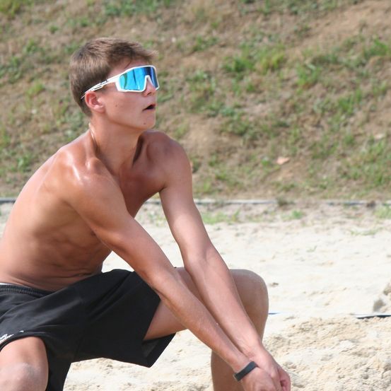 Ein junger Mann ist am Strand, trägt eine Sonnenbrille und schwarze Shorts, mit ausgestreckten Armen und kniet auf dem Sand.