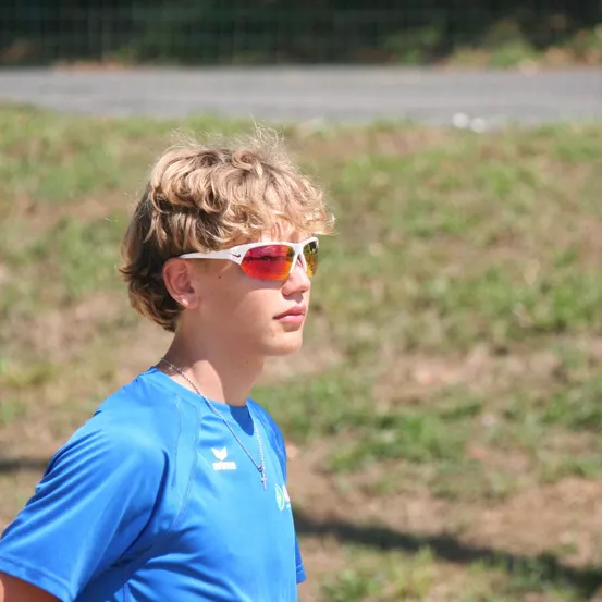 Ein junger Mann in einem blauen Shirt mit Logo steht draußen, trägt eine Sonnenbrille und eine Halskette. Er schaut nach rechts, mit einem verschwommenen Grasbereich hinter ihm.