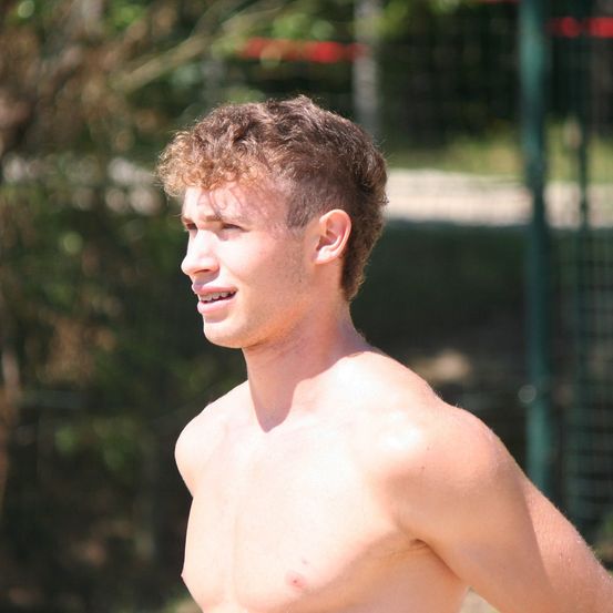 Ein Mann mit lockigem Haar und einem Lächeln steht draußen, ohne Hemd. Bäume und ein Zaun sind im Hintergrund.