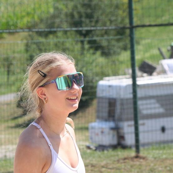 Eine Frau mit Sonnenbrille und einem weißen Tanktop steht draußen und lächelt. Hinter ihr ist ein Zaun mit Grünpflanzen und ein weißer Anhänger.