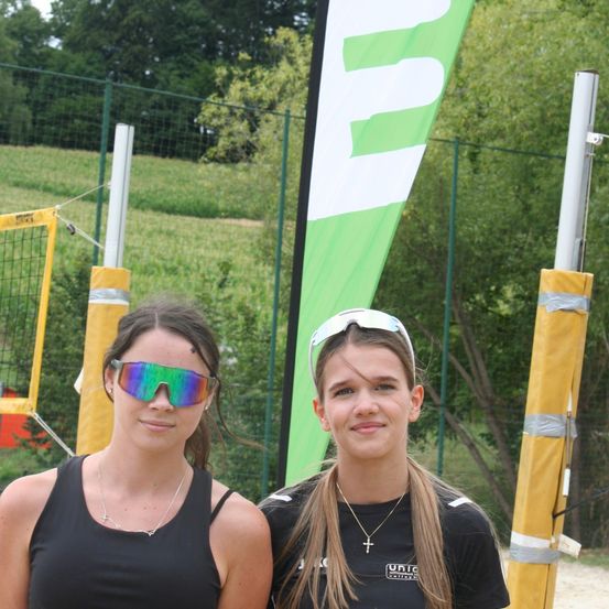 Zwei Frauen stehen vor einem grünen Banner mit weißen Buchstaben. Sie tragen schwarze Tanktops und Sonnenbrillen. Hinter ihnen ist ein Volleyballnetz.