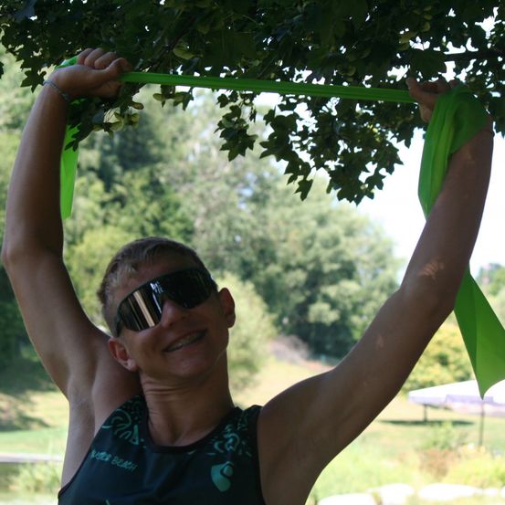 Eine lächelnde Person hält ein grünes Band, das an einem Ast gebunden ist, mit beiden Händen. Die Person trägt eine Sonnenbrille und ein schwarzes Shirt. Im Hintergrund befinden sich Bäume, ein Grasbereich und ein weißes Sonnendach.