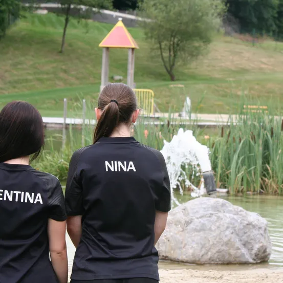 Zwei Frauen, eine mit einem Shirt, auf dem 'Nina' steht, und die andere mit 'Ntina', stehen in der Nähe eines Teiches. Ein Brunnen sprüht Wasser hinter ihnen. Im Hintergrund befindet sich eine Grasfläche und eine kleine Struktur.