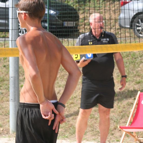 Ein junger Mann steht mit dem Rücken zur Kamera in der Nähe eines Volleyballnetzes. Ein älterer Mann mit Brille und Mikrofon steht in der Nähe. Autos sind hinter dem Netz geparkt.
