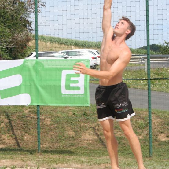 Ein Mann, der Beachvolleyball spielt, streckt seinen Arm hinter sich. Er trägt schwarze Shorts mit weißen Rändern und hat ein Banner mit dem Buchstaben G hinter sich.