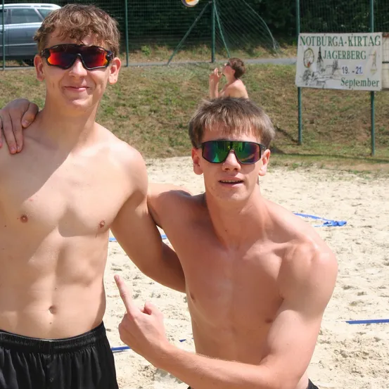 Zwei junge Männer posieren für ein Foto am Strand, beide tragen Sonnenbrillen. Im Hintergrund ist eine Person zu sehen, und ein Schild trägt den Text 'Notburga-Kirtaq Jagerberg' mit einem Datumsbereich.