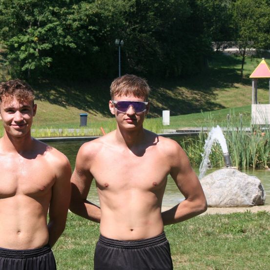 Zwei oberkörperfreie junge Männer stehen in einem Park in der Nähe eines Brunnens und lächeln für die Kamera. Sie tragen beide schwarze Shorts und einer hat eine Sonnenbrille auf.