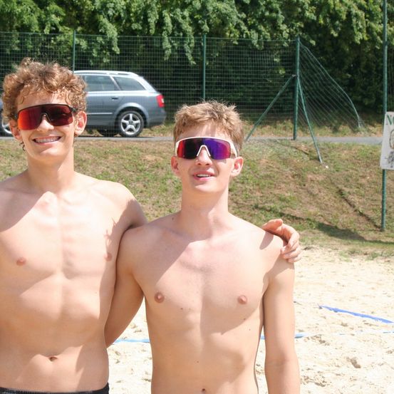 Zwei junge Männer stehen zusammen am Strand, beide tragen Sonnenbrillen. Sie scheinen für ein Foto zu posieren. Hinter ihnen befindet sich ein Grasbereich mit einem Zaun und einem geparkten Auto.