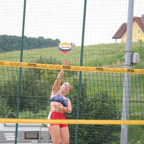 Eine Frau spielt Volleyball, greift nach dem Ball und schlägt ihn, mit einem Netz und einem Haus im Hintergrund.