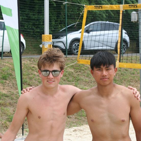Zwei junge Männer stehen am Strand, einer mit dem Arm um die Schulter des anderen. Sie tragen beide eine Sonnenbrille. Hinter ihnen steht ein Beachvolleyballnetz. Ein Auto ist in der Nähe geparkt.