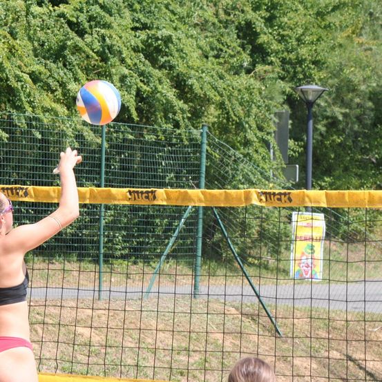 Eine Frau in einem Bikini serviert einen Volleyball über ein Netz, auf dem 'SUCK' steht, während eine andere Person von unten zusieht. Die Szene ist im Freien mit einem grünen Zaun und Bäumen im Hintergrund.