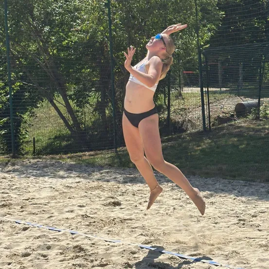 Eine Frau in einem weißen Bikini springt auf einem Sandstrand, möglicherweise Volleyball spielend, mit ausgestreckten Armen.
