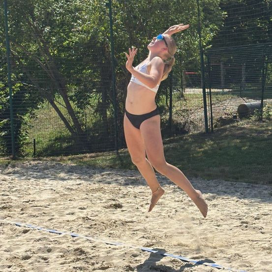 Eine Frau in einem weißen Bikini springt auf einem Sandstrand, möglicherweise Volleyball spielend, mit ausgestreckten Armen.