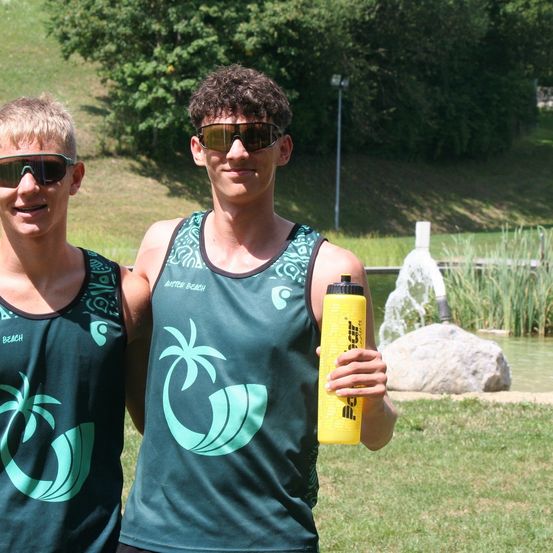 Zwei junge Männer in Sonnenbrillen und ärmellosen Shirts stehen nebeneinander. Einer hält eine gelbe Wasserflasche. Dahinter befindet sich eine Rasenfläche mit einem Felsen und einer Wasserfontäne. Im Hintergrund sind Bäume und ein Laternenpfahl zu sehen.