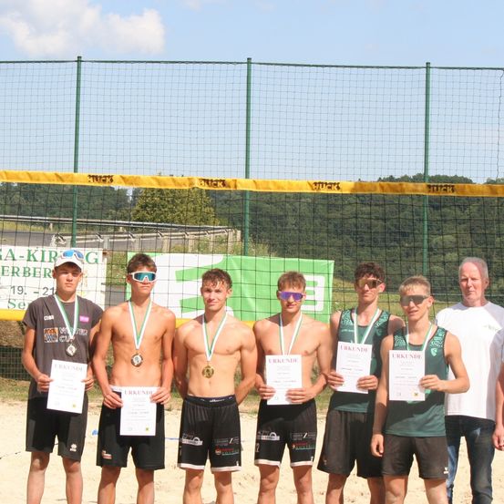 Eine Gruppe junger Männer mit Medaillen und Urkunden steht am Strand und posiert für ein Foto. Hinter ihnen befindet sich ein grünes Netz.