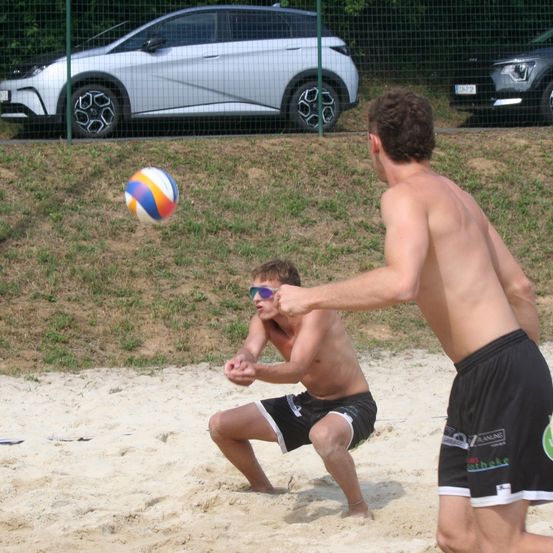 Zwei Männer spielen Beachvolleyball auf einem sandigen Feld, hinter ihnen ist ein Auto hinter einem Zaun geparkt.