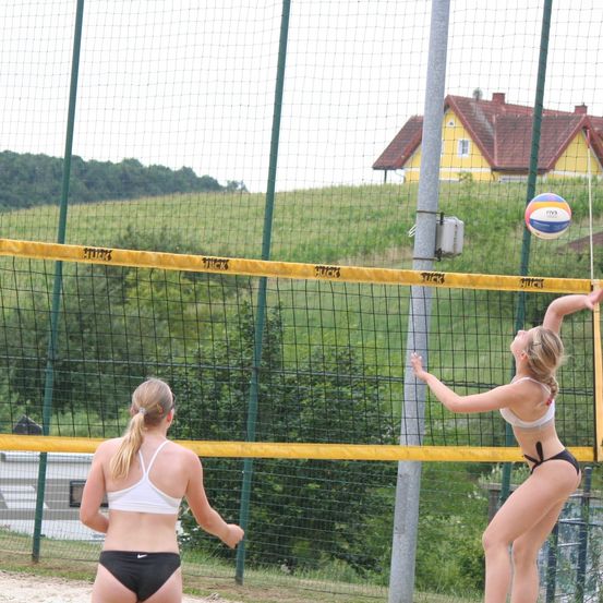 Zwei Frauen spielen Volleyball. Eine Spielerin springt, um den Ball zu schlagen, während die andere den Ball beobachtet. Ein Haus ist im Hintergrund.