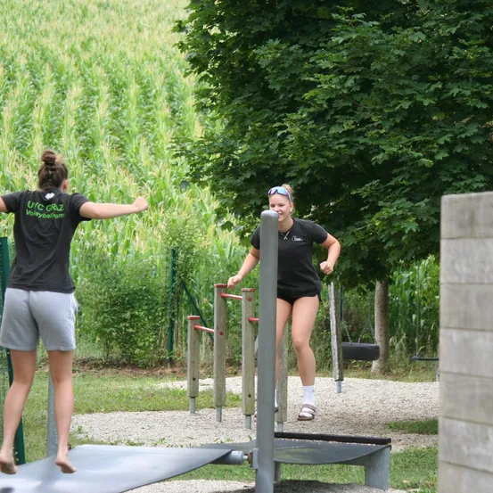 Zwei Frauen trainieren in einem Outdoor-Fitnessstudio. Eine steht auf einem Balancebrett, die andere läuft auf einem Laufband. Sie tragen schwarze T-Shirts und Shorts. Hinter ihnen steht ein Baum und eine Reifenschaukel.