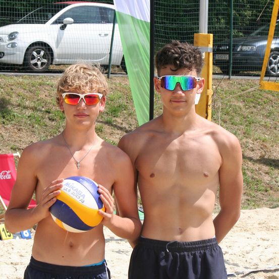 Zwei junge Männer am Strand, einer hält einen Volleyball, beide tragen Sonnenbrillen, mit einem roten Stuhl und einer Fahne im Hintergrund.