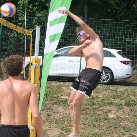 Zwei Männer spielen Beachvolleyball, einer springt, um den Ball zu schlagen. Hinter ihnen steht eine grün-weiße Fahne, und ein weißer Wagen ist außerhalb des Zauns geparkt.