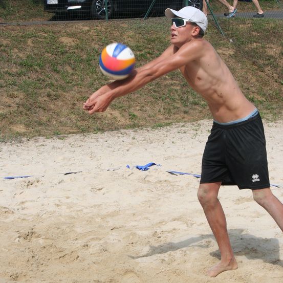 Ein junger Mann in einer weißen Kappe und Sonnenbrille spielt Beachvolleyball. Er ist dabei, den Ball zu schlagen. Hinter ihm befindet sich ein Zaun und einige gehende Personen.