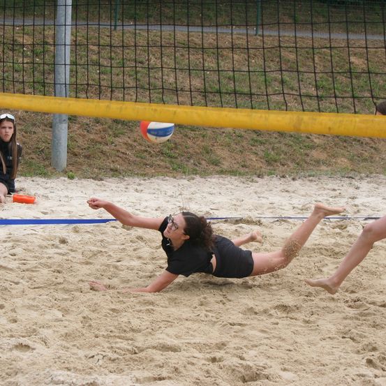 Eine Volleyballspielerin taucht im Sand, um den Ball in der Nähe des Netzes zu retten, während eine andere Spielerin in der Nähe sitzt.